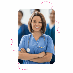 A care provider smiling whilst wearing blue scrubs. Theri arms are folded. The background is blurred slightly but there are a number of additional care providerds standing in the background.