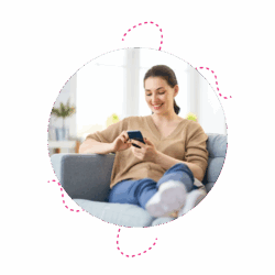 A woman sitting on a grey couch smiling at her phone. The background is blured but it is evident she is in her own home.