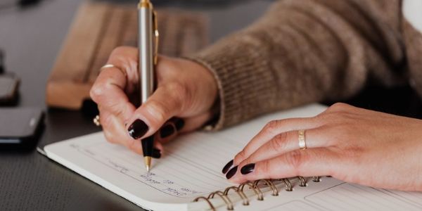 Close-up of a woman’s hands writing notes with a pen in a spiral notebook, planning care or keeping a journal at a desk.